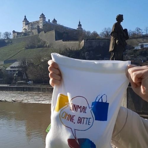 Foto eines hochgehaltenen Stoffbeutels mit Logo von „Einmal ohne, bitte“ vor der Kulisse Würzburgs