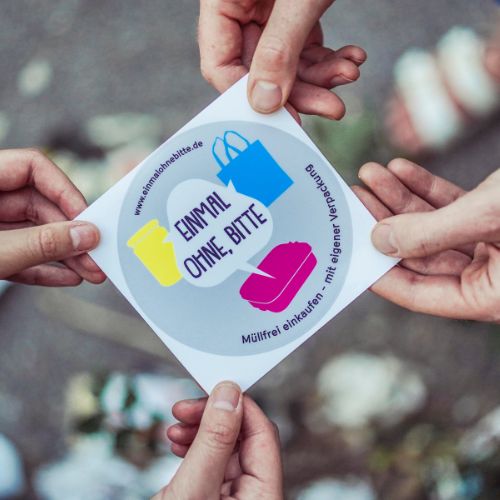 Vier Hände halten den Sticker von „Einmal ohne, bitte“ an seinen Ecken