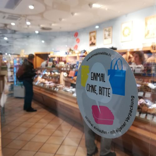 Foto durchs Schaufenster mit Sticker von „Einmal ohne, bitte“ in ein Lebensmittelgeschäft