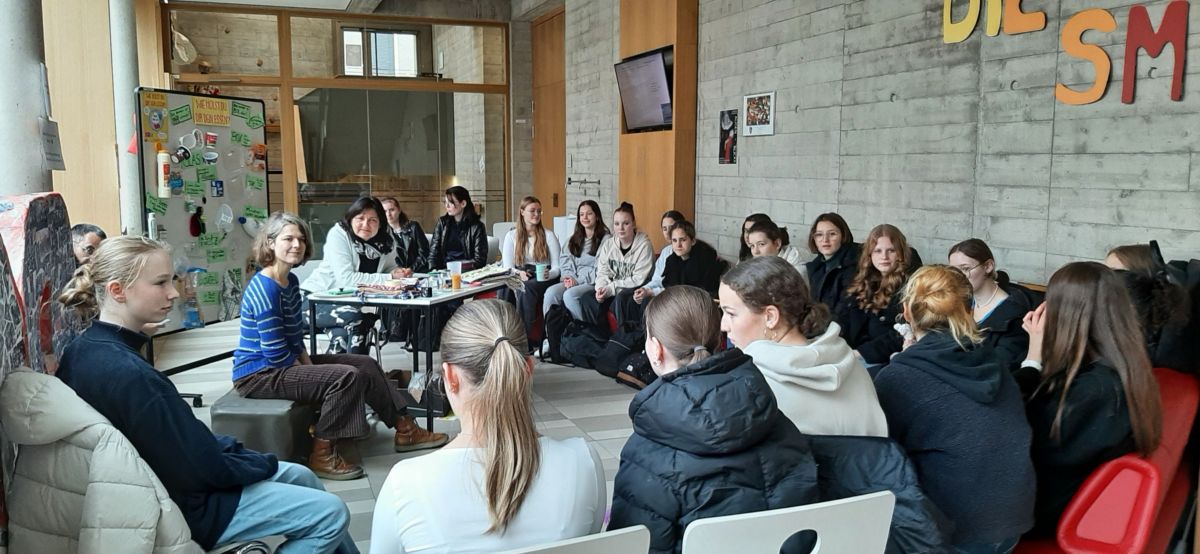 Foto einer Schulklasse im Stuhlkreis; zwei Workshop-Leiterinnen sitzen vor einem Schaubild