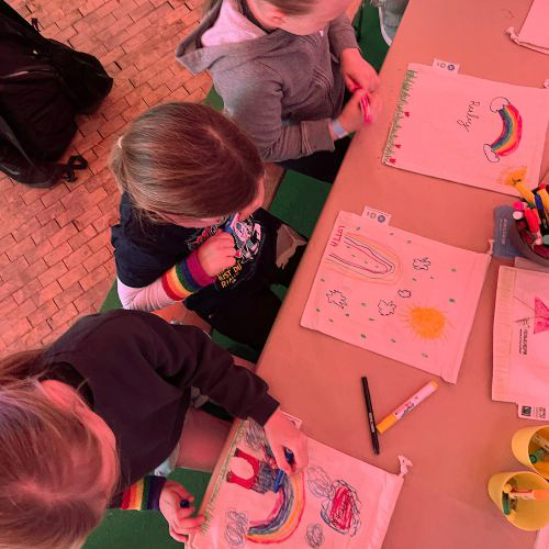 Foto einer Gruppe Kinder, die rund um einen Tisch stehend Stoffbeutel mit Namen, Begriffen und Symbolen bemalen; in der Mitte steht ein Becher mit Textilstiften