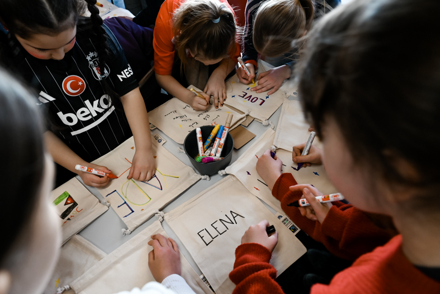 Foto von drei Kindern, die an einem Tisch sitzen und mit Textilstiften ihren Namen und Regenbogen-Motive auf Stoffbeutel malen