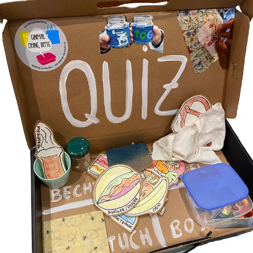 Foto eines Pappkoffers mit der Aufschrift „Quiz“, gebastelten Lebenssymbolen und Mehrwegbehältnissen