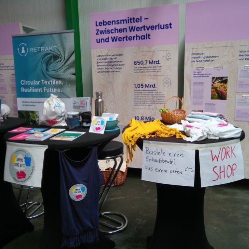 Foto eines Infostands von „Einmal ohne, bitte“ mit einer Bastelstation. Auf zwei Schildern steht: „Workshop“ und „Bastle einen Einkaufsbeutel aus einem alten T-Shirt“.
