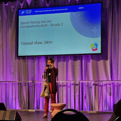 Foto einer Person, die auf der Bühne einen Pitch zu „Einmal ohne, bitte“ hält. Auf dem Bildschirm im Hintergrund steht: „Speed-Dating mit der Kreislaufwirtschaft“.